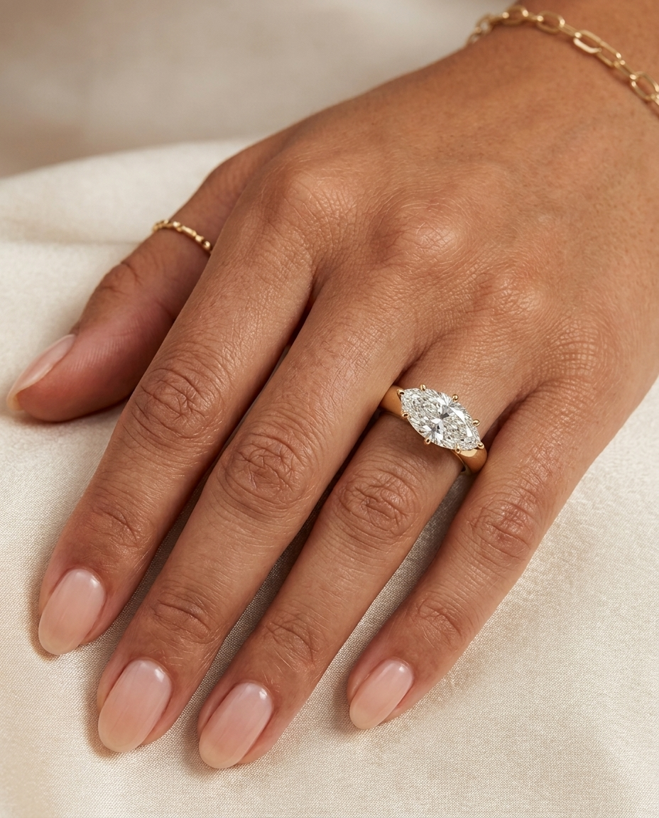 3ct east west marquise diamond solitaire A close-up of a hand wearing a yellow gold ring with a marquise-cut clear gemstone, resting on a soft cream fabric background.