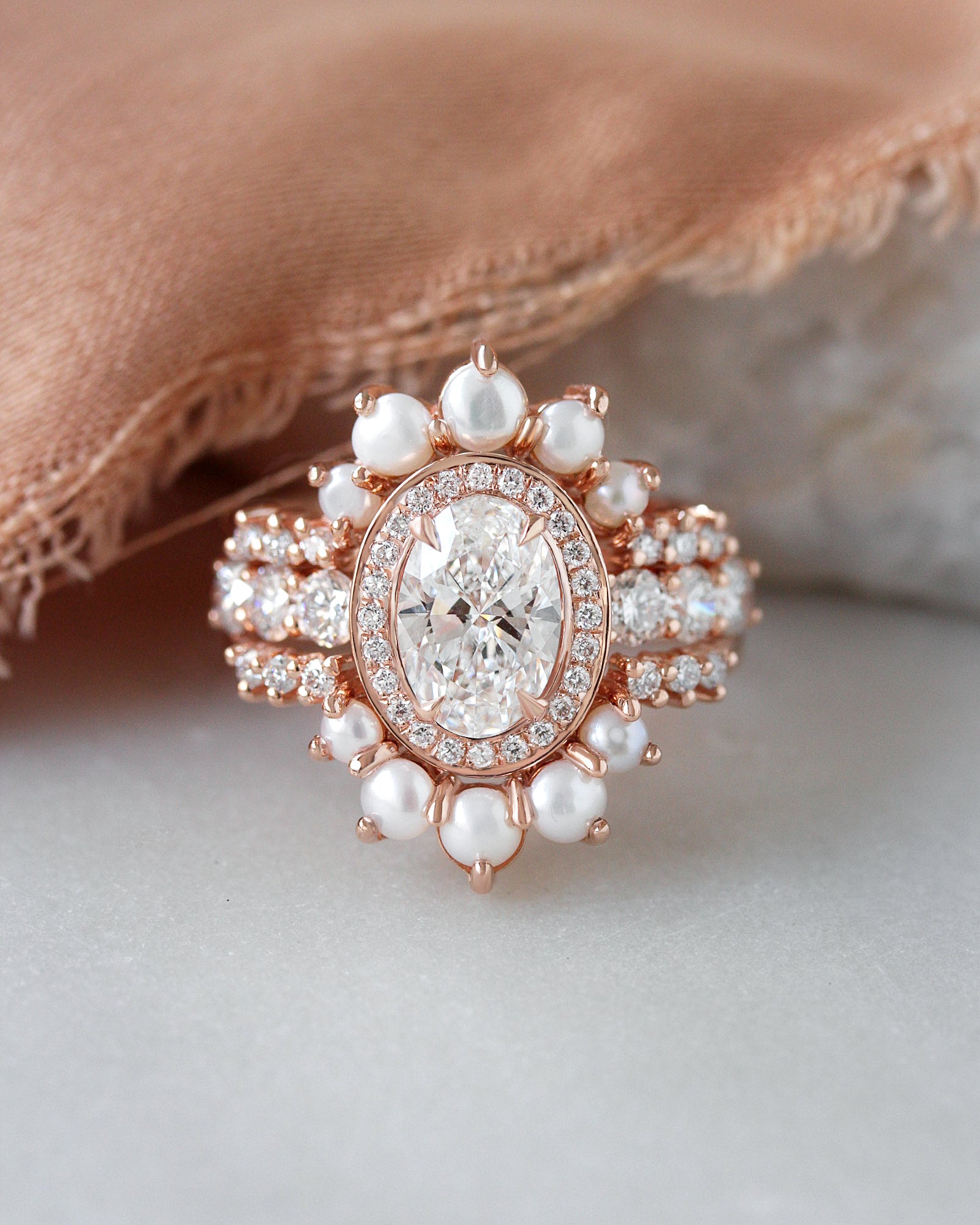 Rose gold halo ring with a large oval center stone and surrounding pearls and small clear stones, photographed on a soft neutral surface with a beige fabric prop.