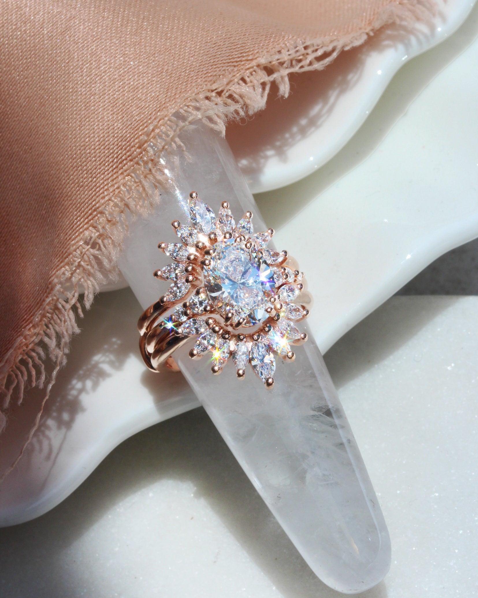 2ct oval diamond engagement ring. 
A rose gold ring with a large clear center stone and surrounding marquise-cut stones, displayed on a clear crystal cone prop over a white surface with a peach fabric drape.
