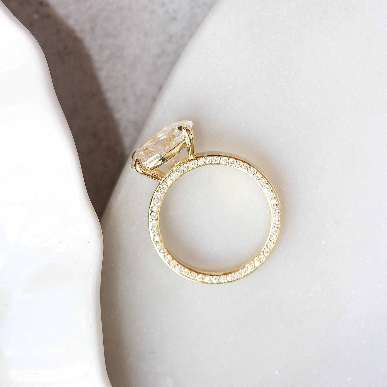3CT OVAL diamond hidden eternity engagement ring A gold ring with a large clear faceted gemstone and a band paved with small clear stones, resting on a white textured surface.