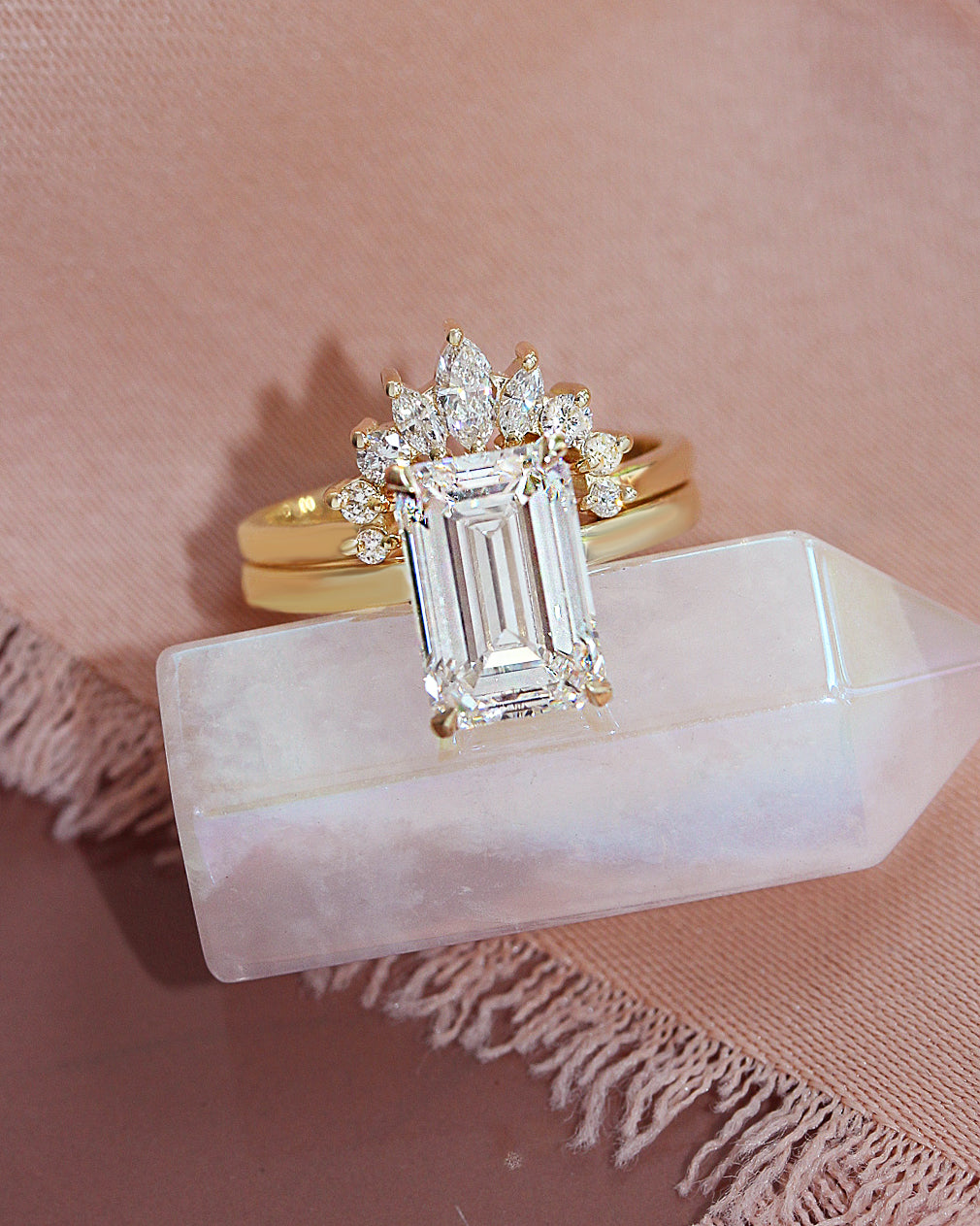 4CT emerald cut diamond Solitaire engagement ring with a matching nesting ring . A gold ring with a large emerald-cut clear center stone and smaller surrounding stones, displayed on a pale pink stone block against a dusty pink fabric background.