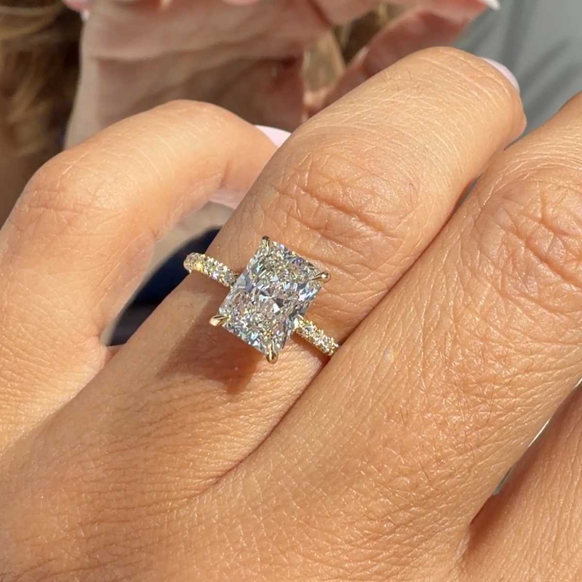 A close-up of a hand wearing a yellow gold ring with a rectangular clear gemstone, with a phone visible in the background.